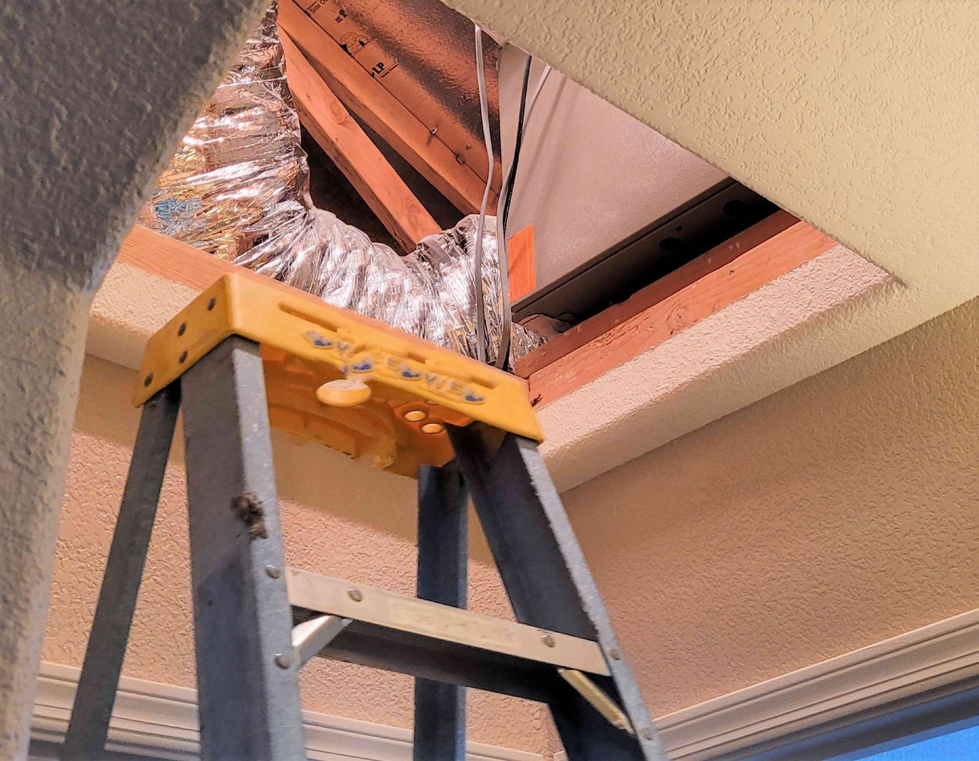 A ladder inside a room, leading to an open ceiling hatch with ductwork in the attic.
