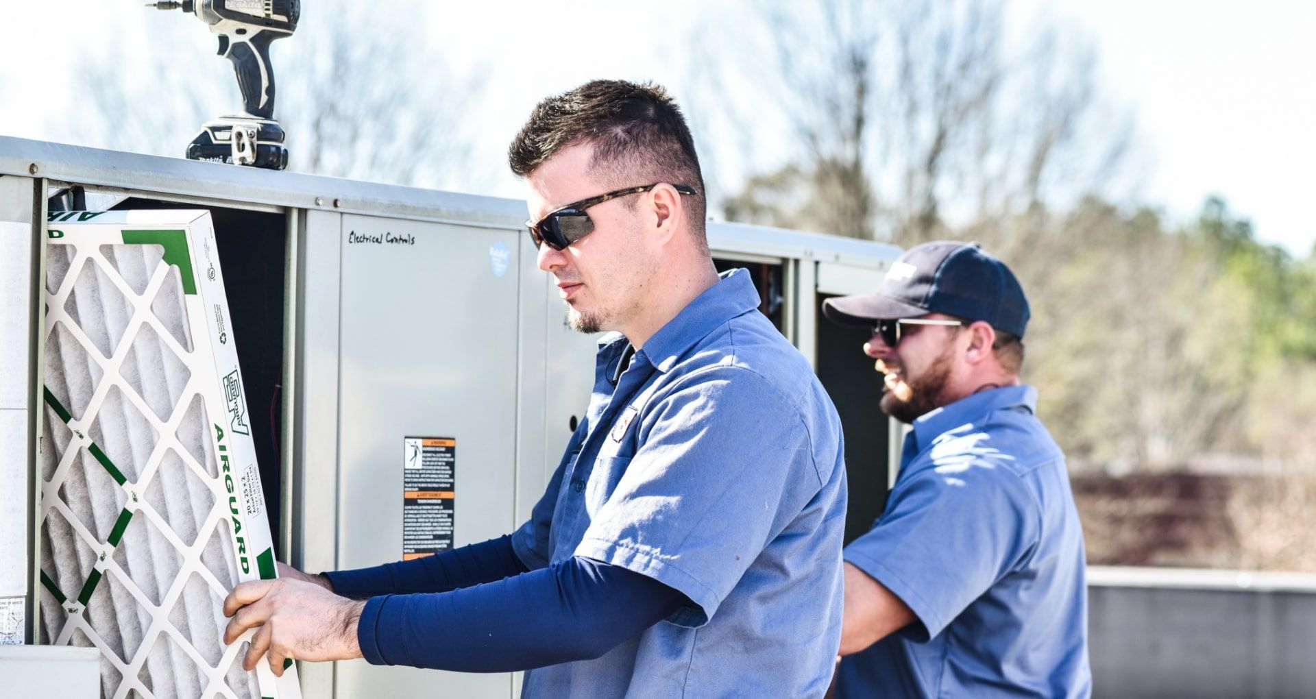 Two men in blue shirts replacing an air filter on a rooftop HVAC unit. Sunny day.