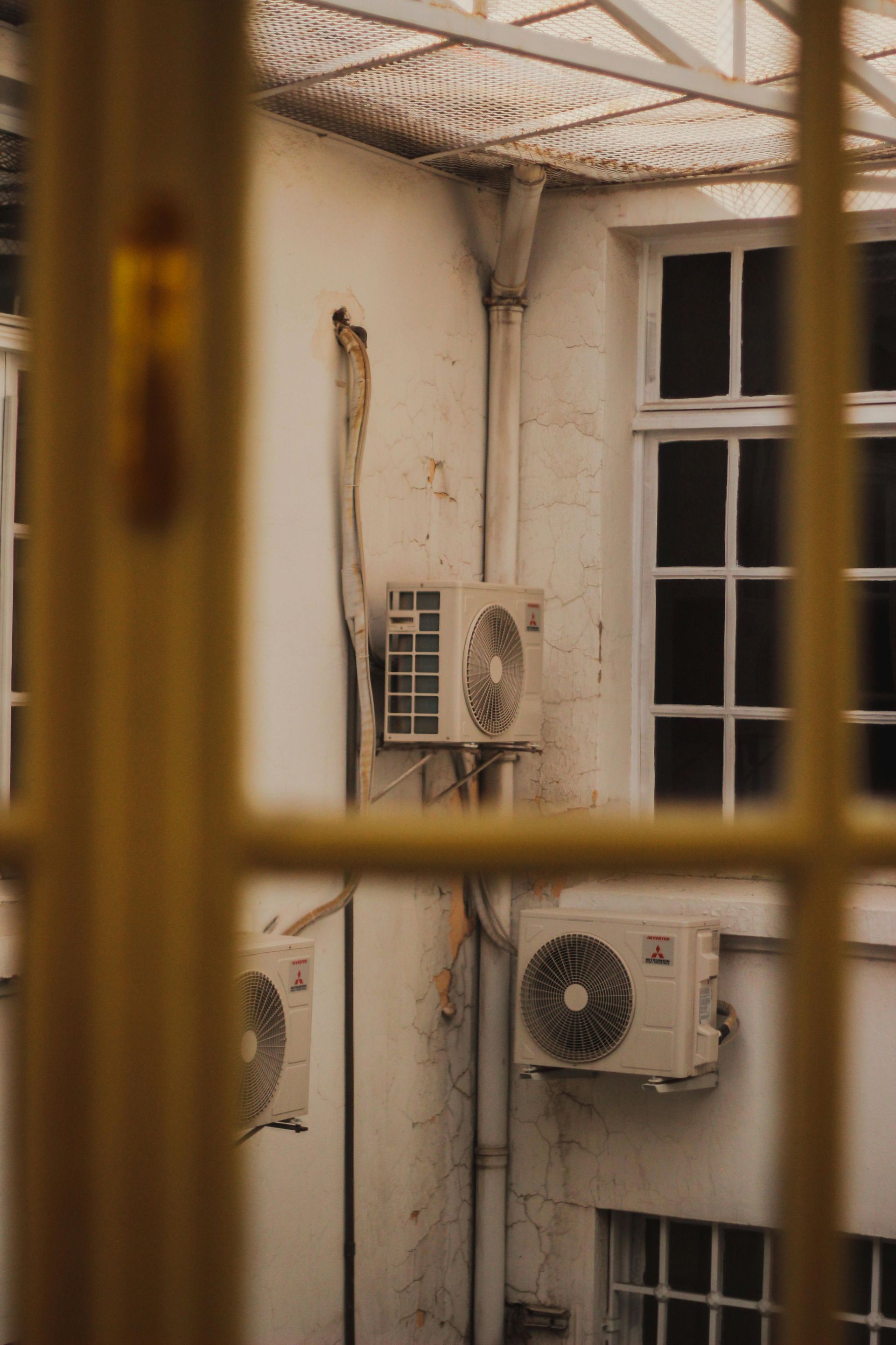 View of a building courtyard with air conditioning units mounted on a white wall, framed by a window.