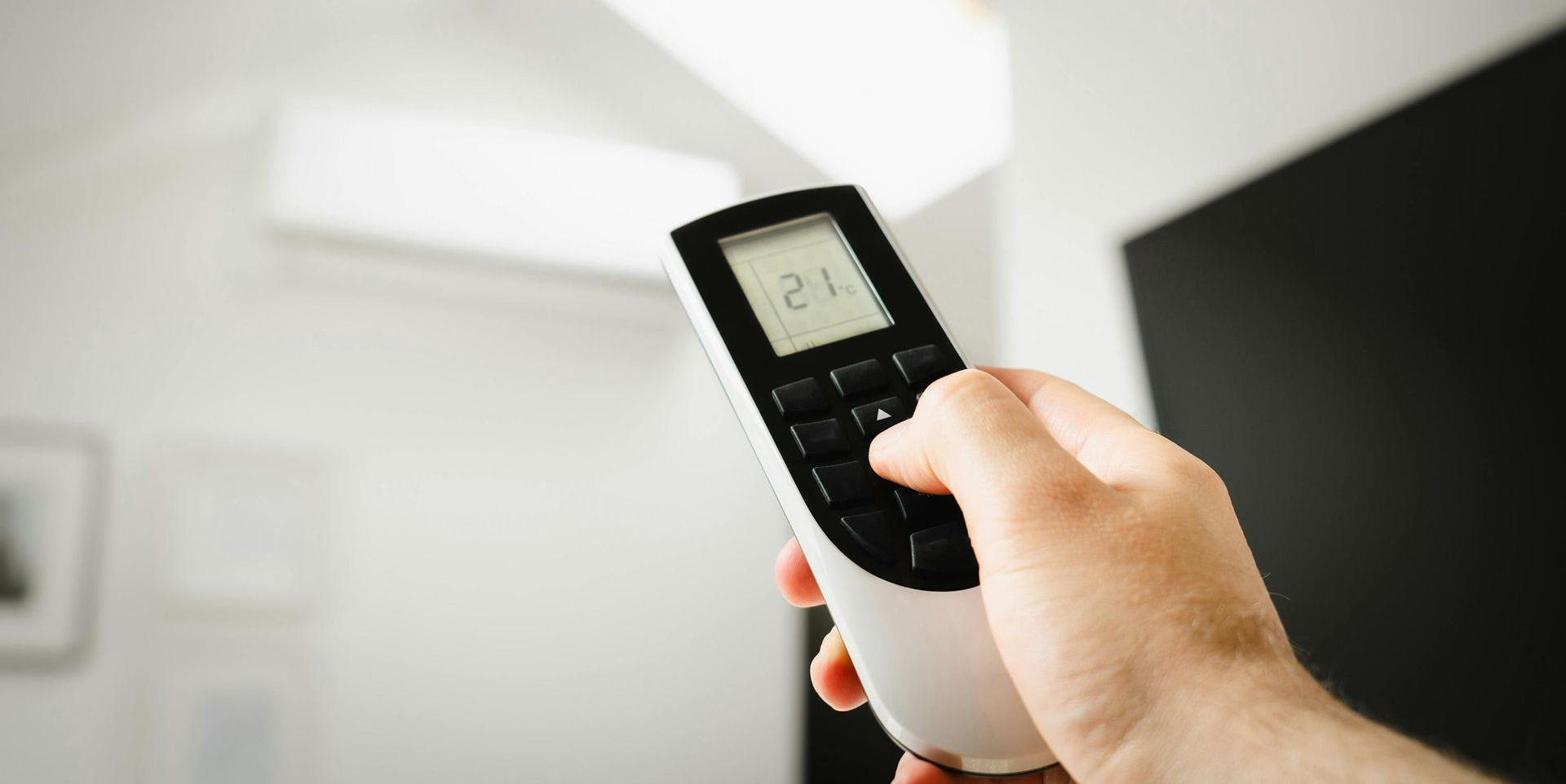 A hand holding an air conditioner remote, pressing a button. An AC unit is in the background.