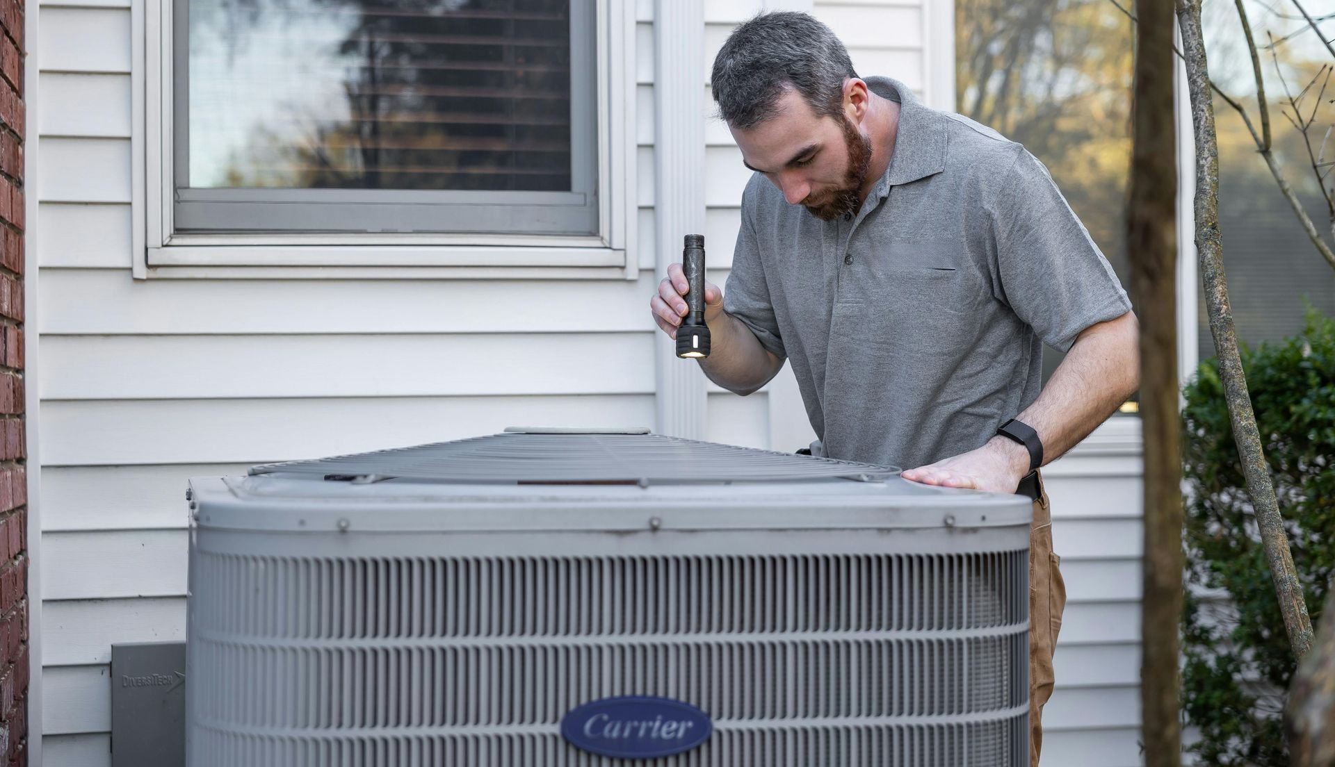 Man inspecting an air conditioning unit outdoors, near a window; he wears a gray shirt, and has a beard.