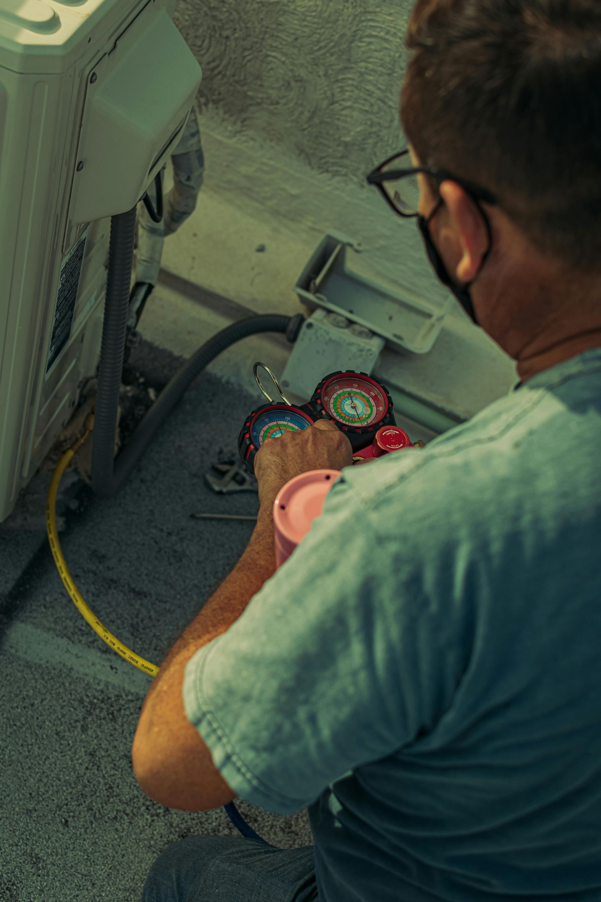 A technician wearing glasses and a mask working on an AC unit outdoors, connecting gauges.