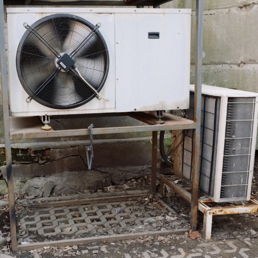 Air conditioning units, a large one and a smaller one, on a metal stand outdoors against a concrete wall.