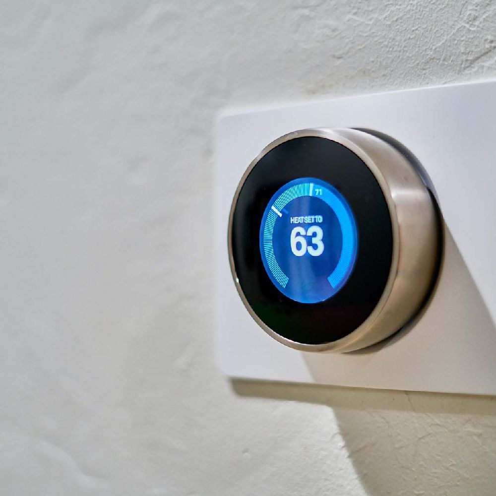 Smart thermostat with blue display showing temperature of 63 degrees. Mounted on white wall.