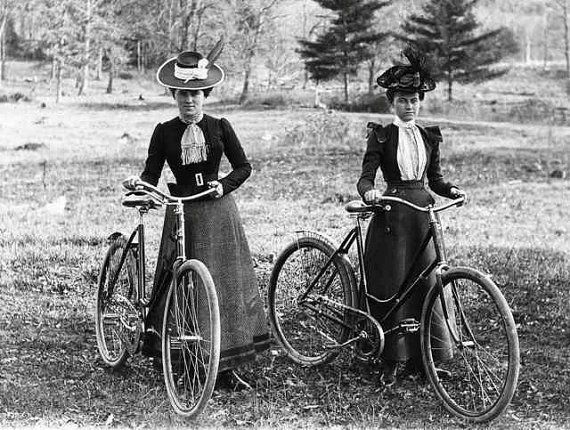 Two women in hats and long skirts stand with bicycles outdoors.