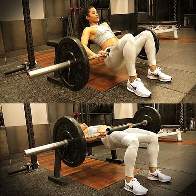 Woman performing barbell hip thrusts in a gym, lifting weight, wearing sportswear, with focused expression.