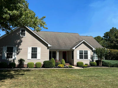 Suburban beige house with a gray roof, white-trim windows, and a manicured lawn under a blue sky