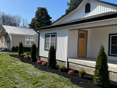 White house with porch, tan front door, and landscaped garden beds under a clear blue sky.
