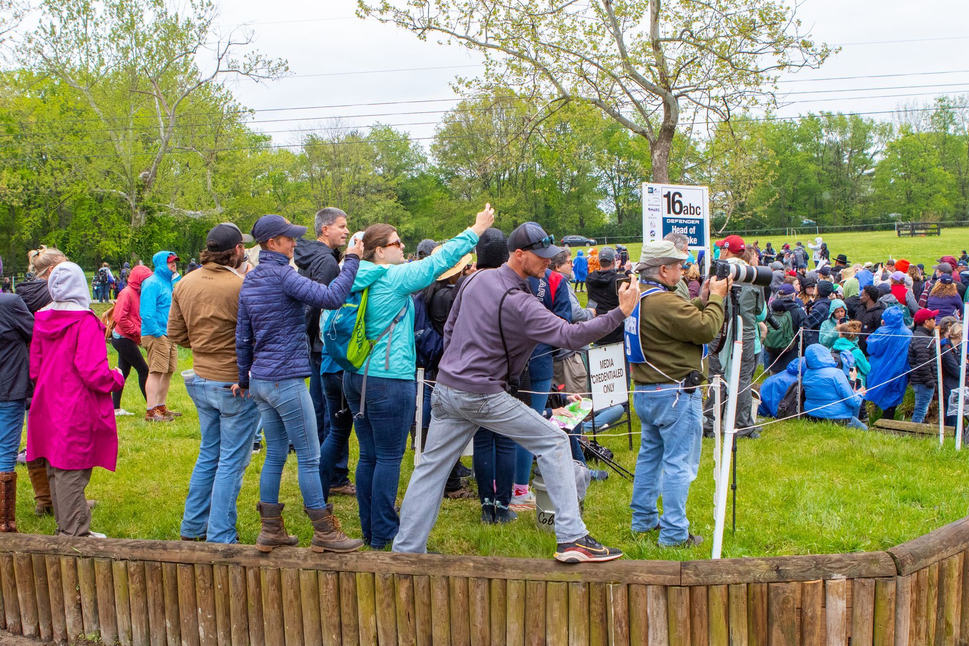 A large group of people are standing around a wooden fence in a park.