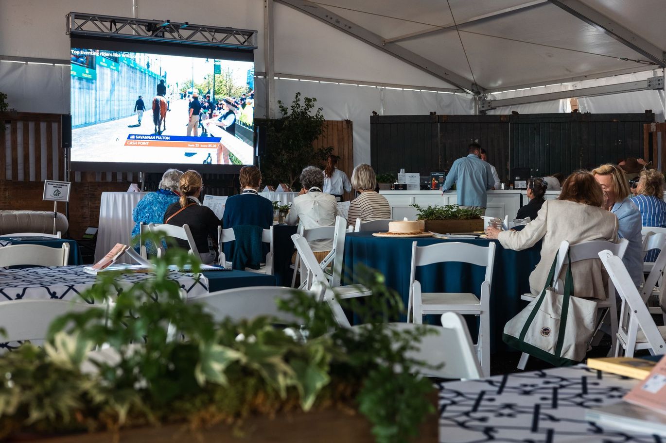 A group of people are sitting at tables in front of a large screen.