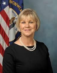 A woman in a black dress and pearl necklace is standing in front of an american flag.