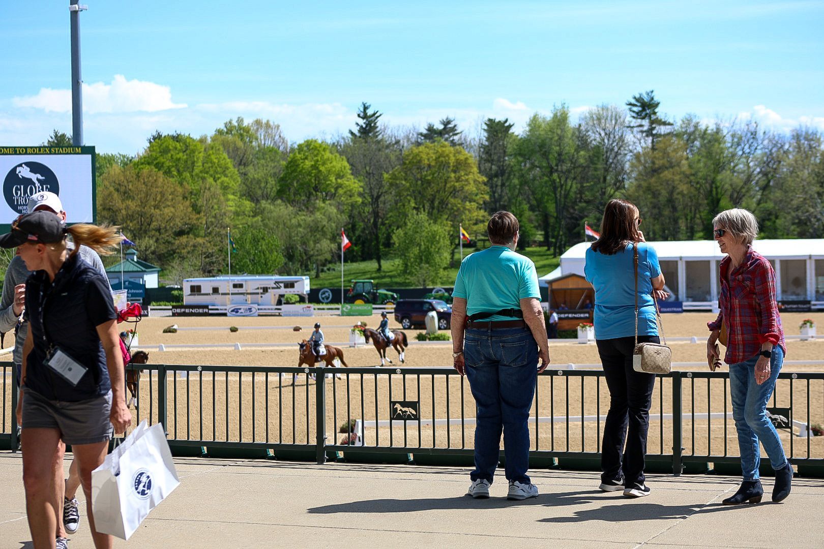 Three women stand overlooking horse and riders in an arena