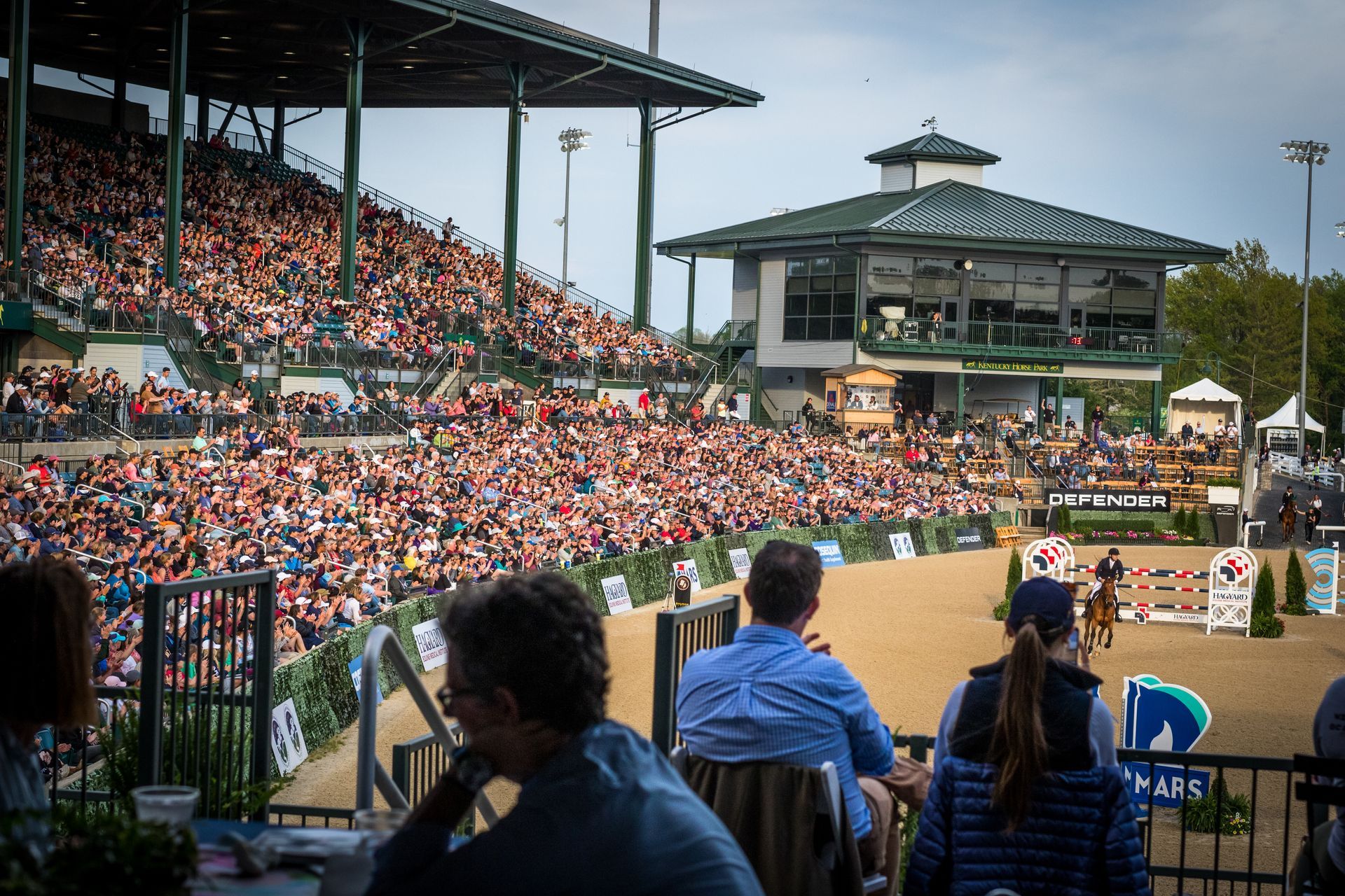 A crowd of people are watching a horse race in a stadium.