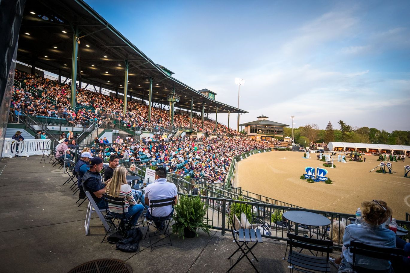 A group of people are sitting at tables in a stadium watching a horse show.