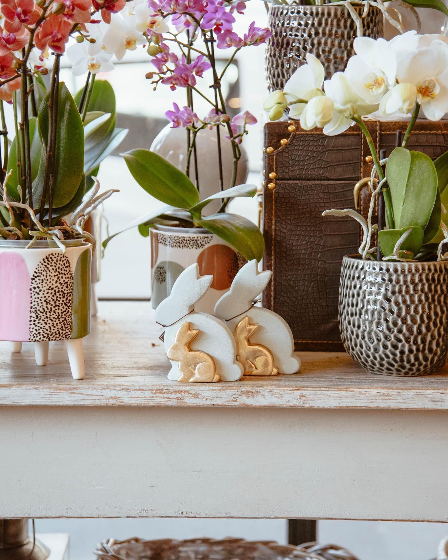 Composizione pasquale con orchidee, decorazioni a forma di coniglietto e vasi su un tavolo bianco.