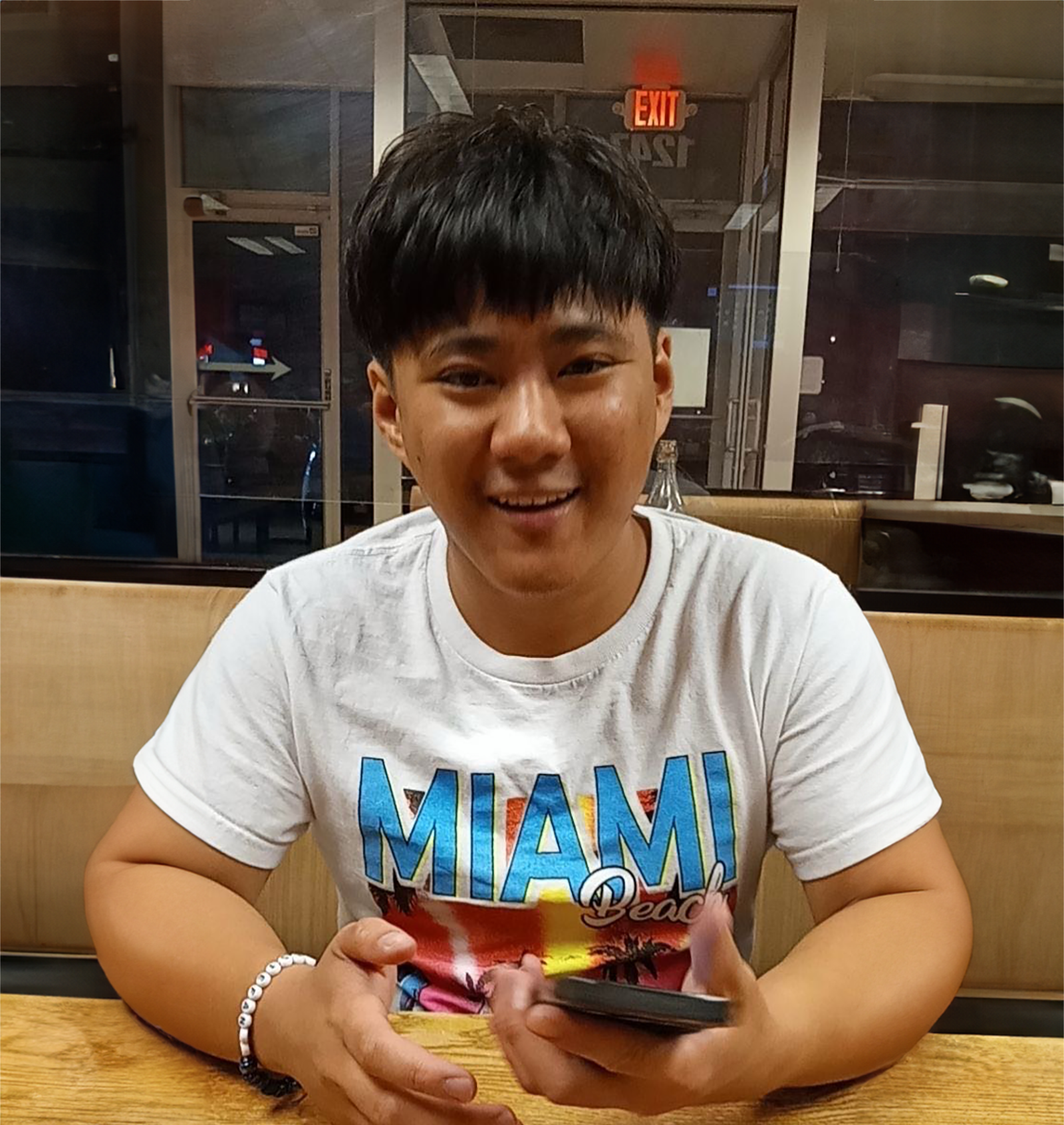 A young man wearing a miami shirt is sitting at a table holding a cell phone
