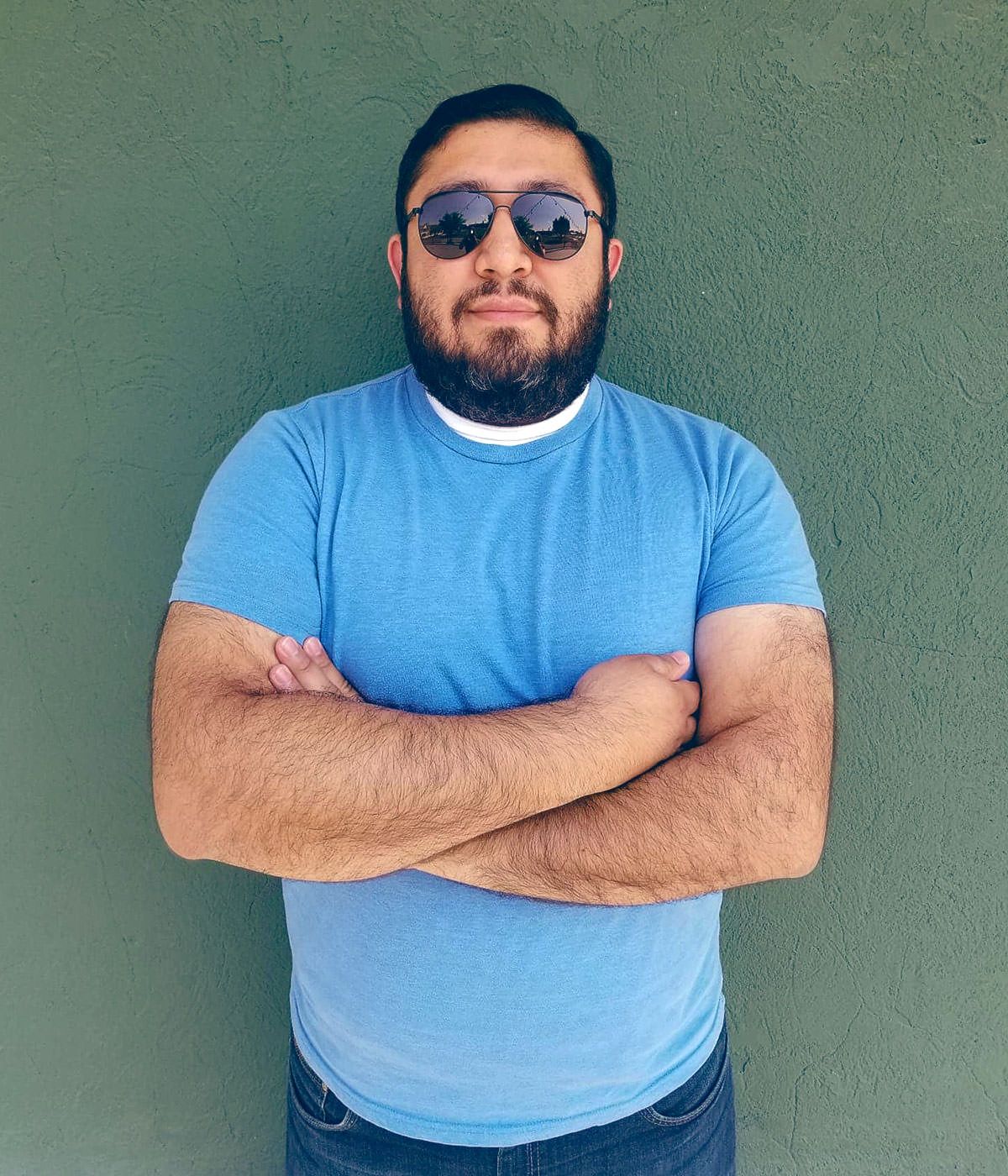 A man with a beard wearing sunglasses and a blue shirt is standing with his arms crossed.