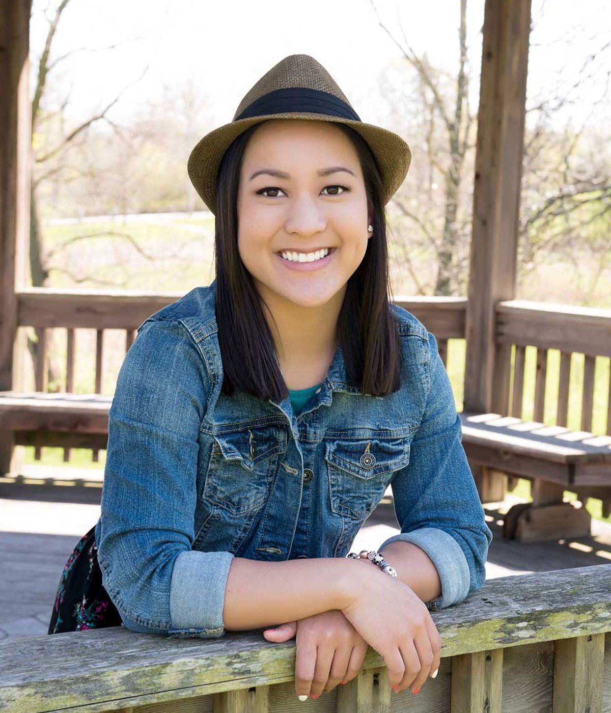 A woman wearing a hat and a denim jacket is sitting on a railing.