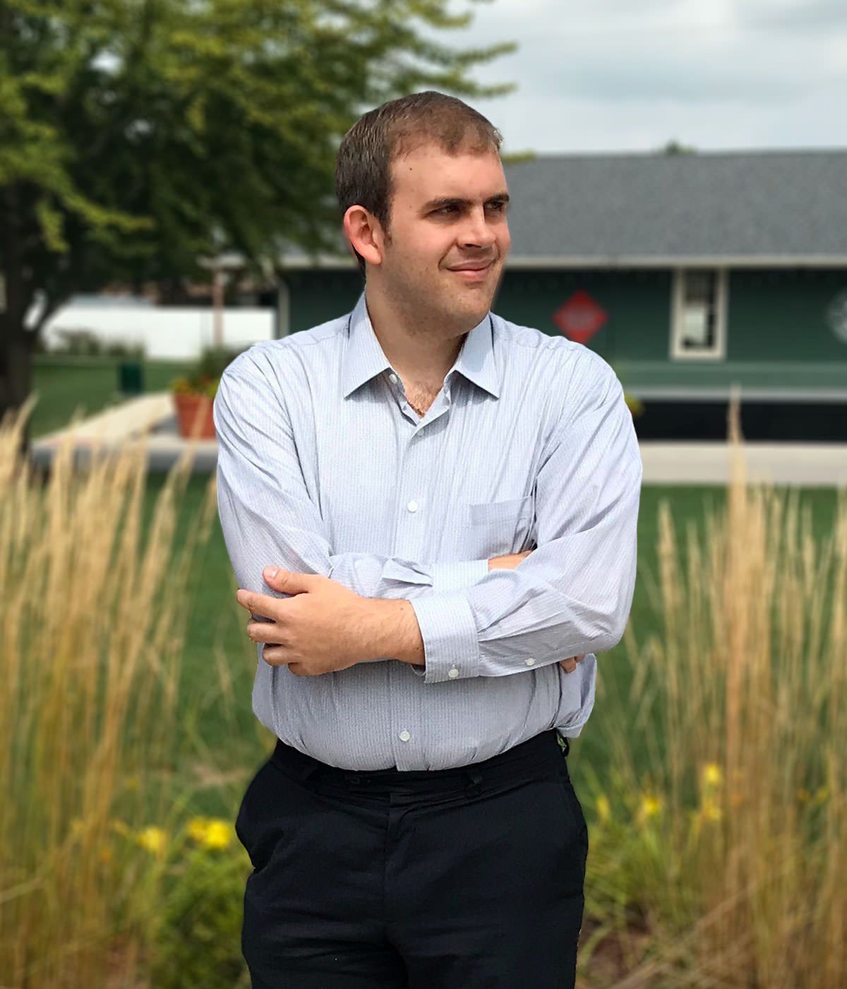 A man in a striped shirt and black pants is standing in front of a house with his arms crossed.
