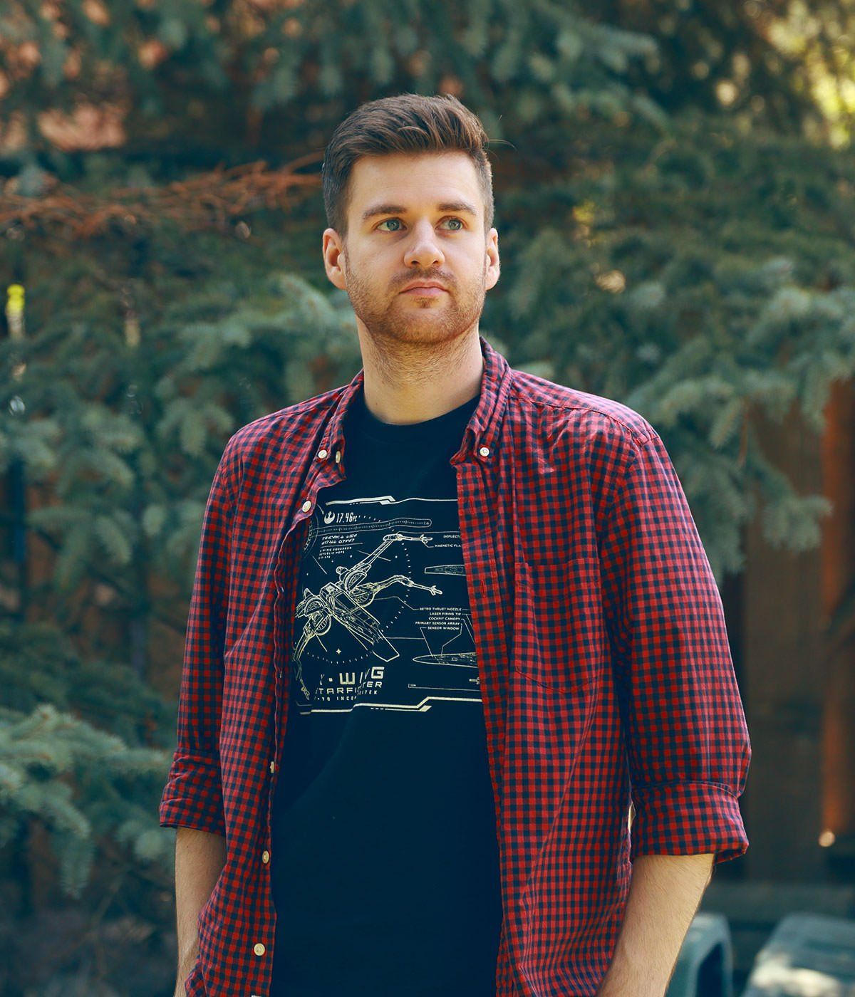A man wearing a plaid shirt and a black t-shirt is standing in front of trees.