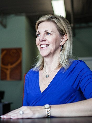 A woman in a blue shirt is sitting at a table and smiling.