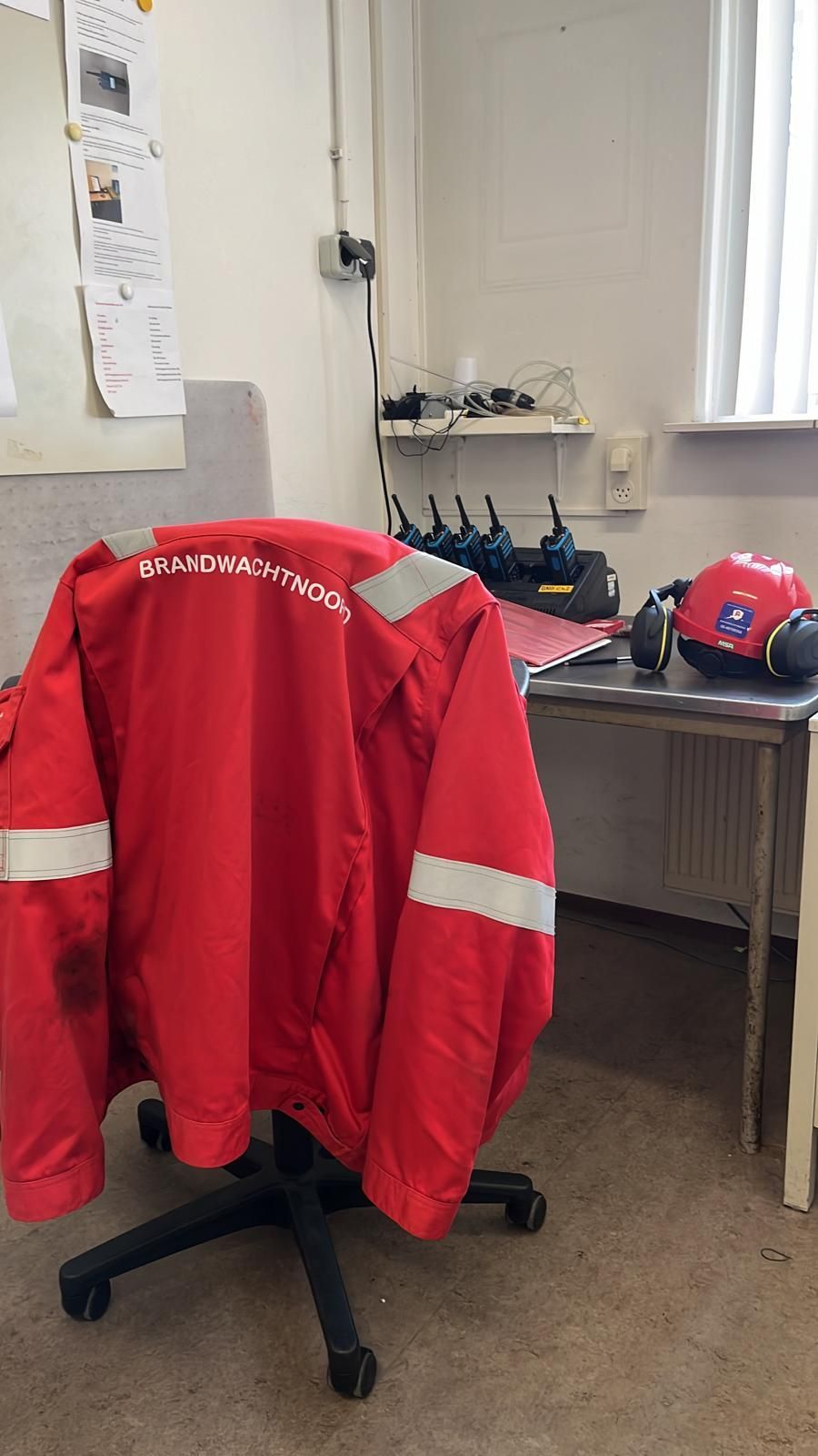 Een rode jas van de hulpdiensten hangt aan een bureaustoel in een kamer met een bureau en apparatuur.