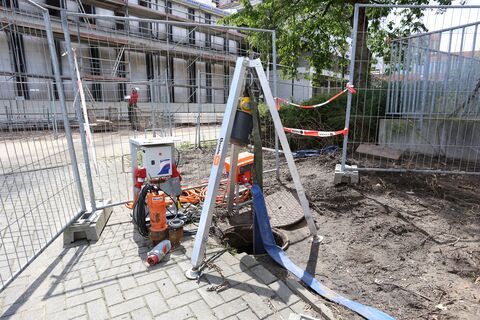Een bouwtripod staat boven een open putdeksel op een verhard oppervlak naast een elektrische pomp en een draadhek.