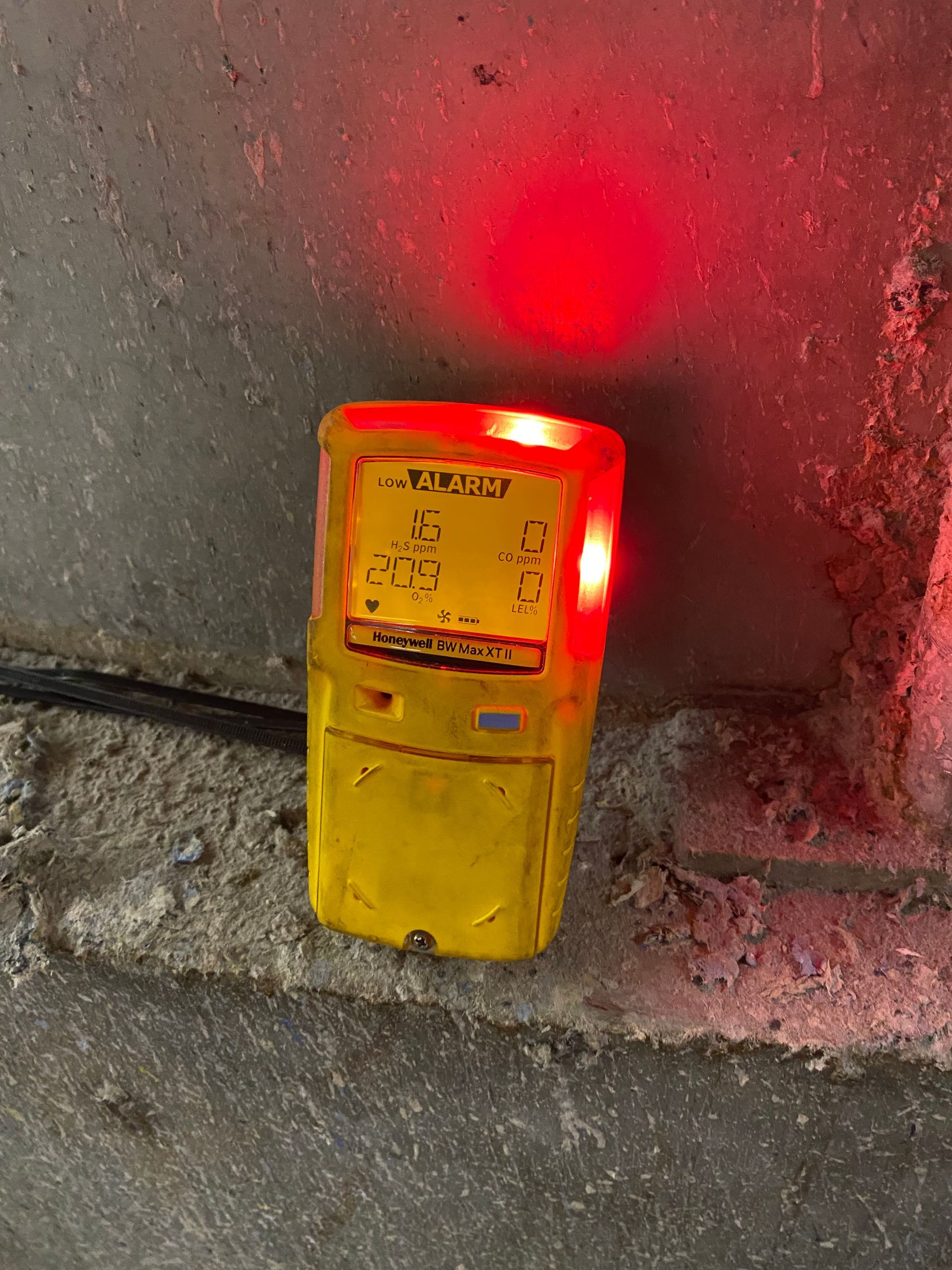 Een gele gasmelder met knipperend rood alarmlicht, geplaatst op een ruwe betonnen vloer vlakbij een muur.