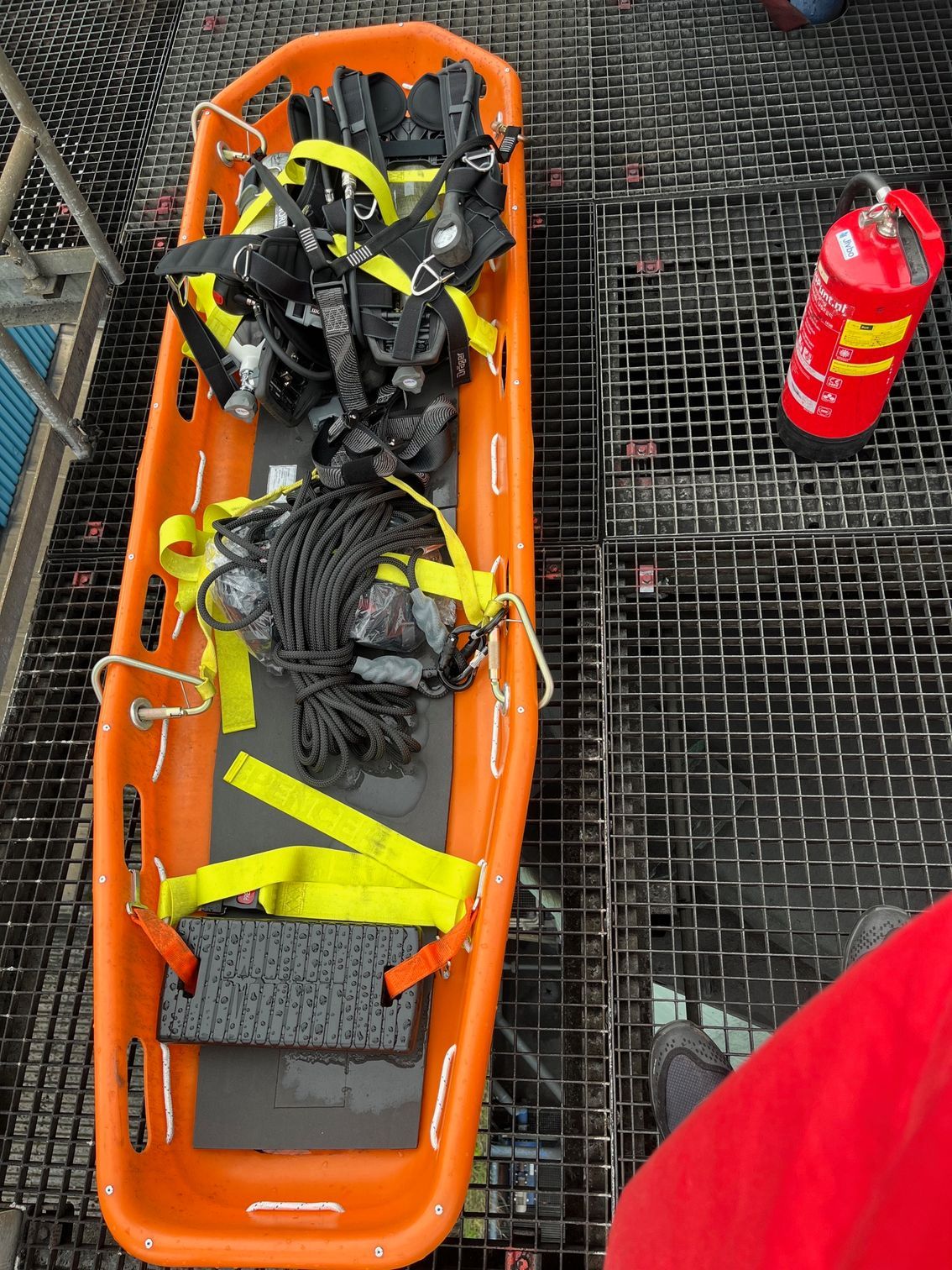 Een oranje reddingsbrancard met veiligheidsriemen en touw, geplaatst op een donker metalen rooster naast een rode brandblusser.