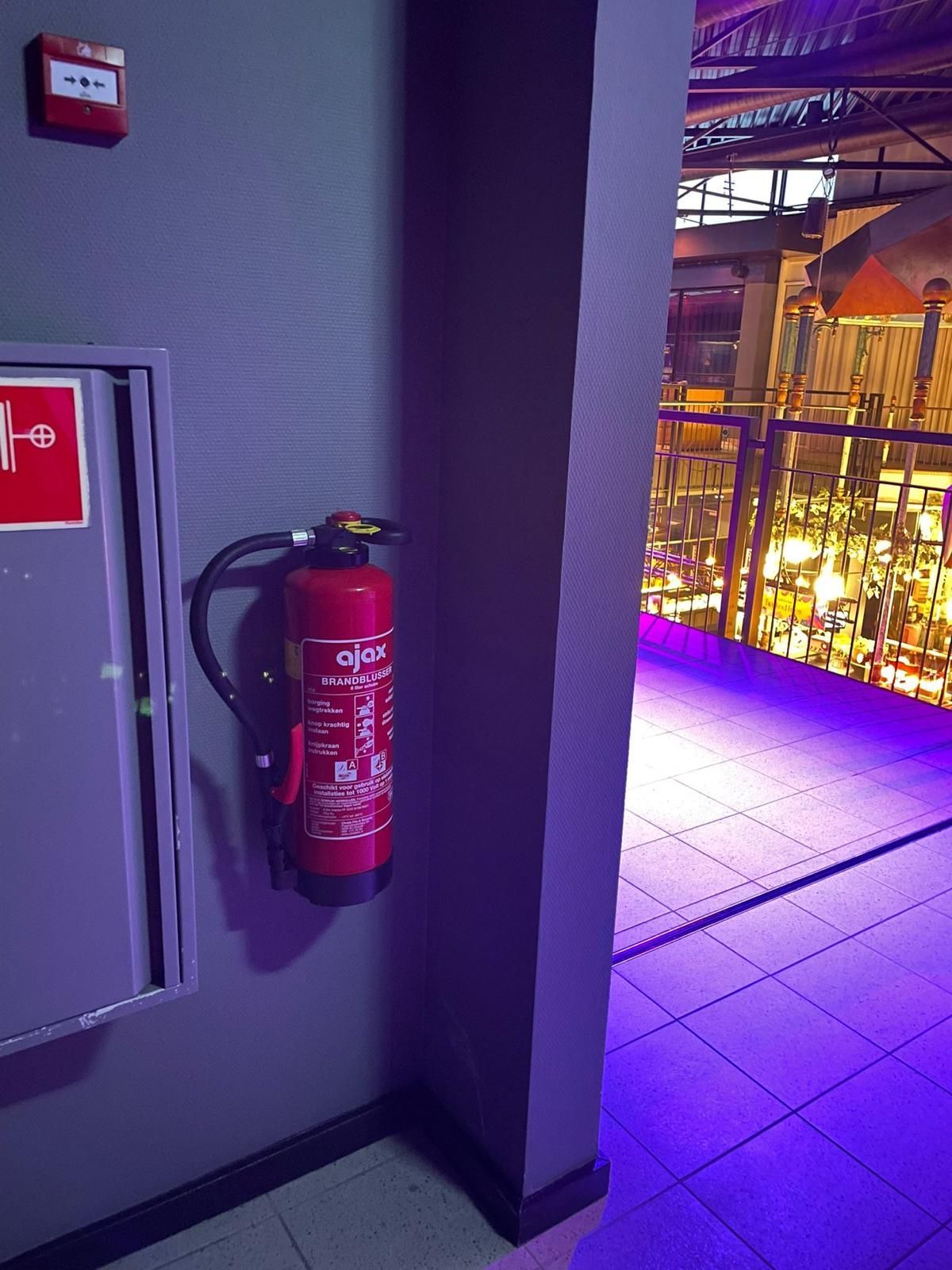 Een rode brandblusser is gemonteerd aan een grijze muur naast een brandalarmknop en een open deuropening naar een balkon.