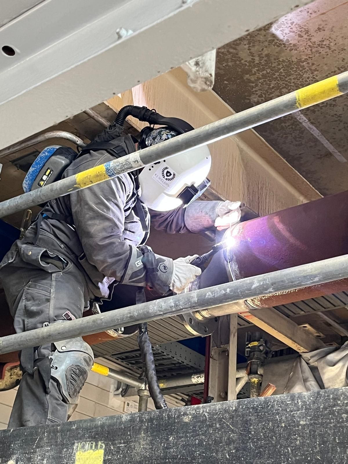 Een lasser, volledig uitgerust met beschermende kleding, werkt aan een grote pijp bovenop een bouwsteiger.