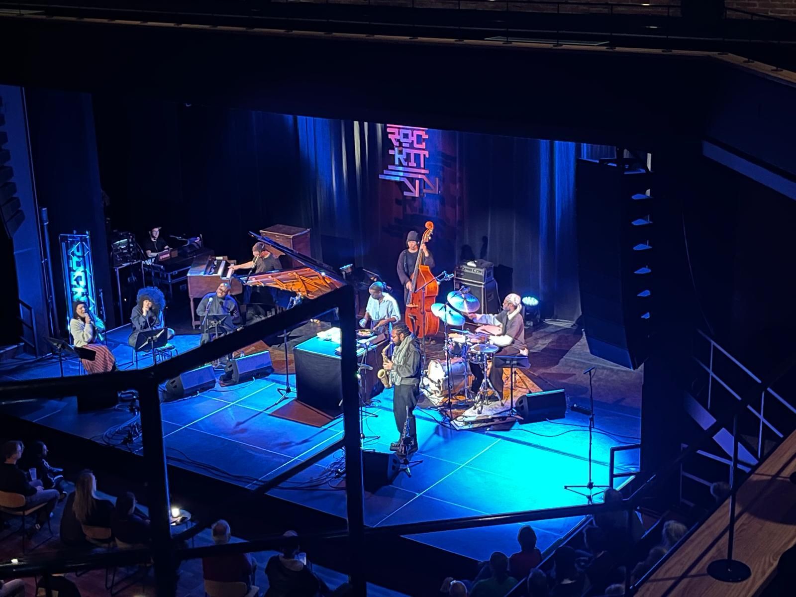 Een jazzband treedt op een fel verlicht blauw podium in een concertzaal, gezien vanuit een verhoogd, maar belemmerd perspectief.