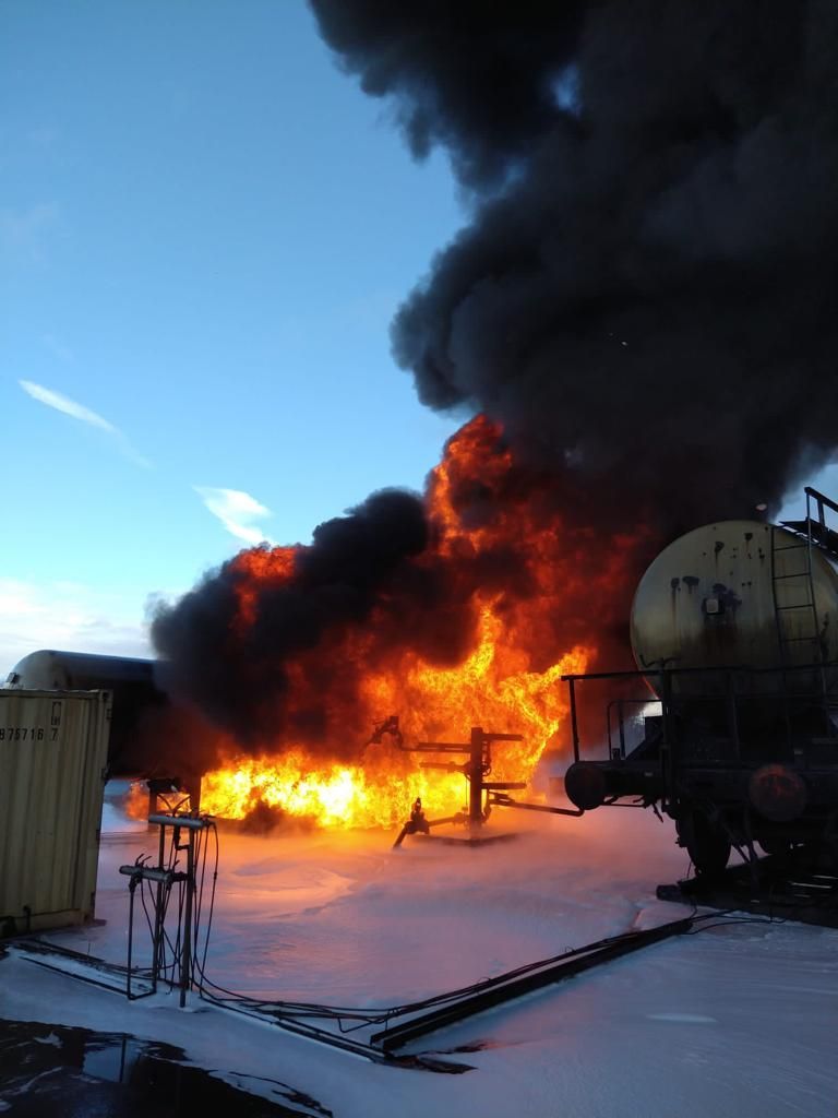 Tussen twee industriële tankwagons woedt een hevige brand, waarbij dikke zwarte rook afsteekt tegen een blauwe hemel.