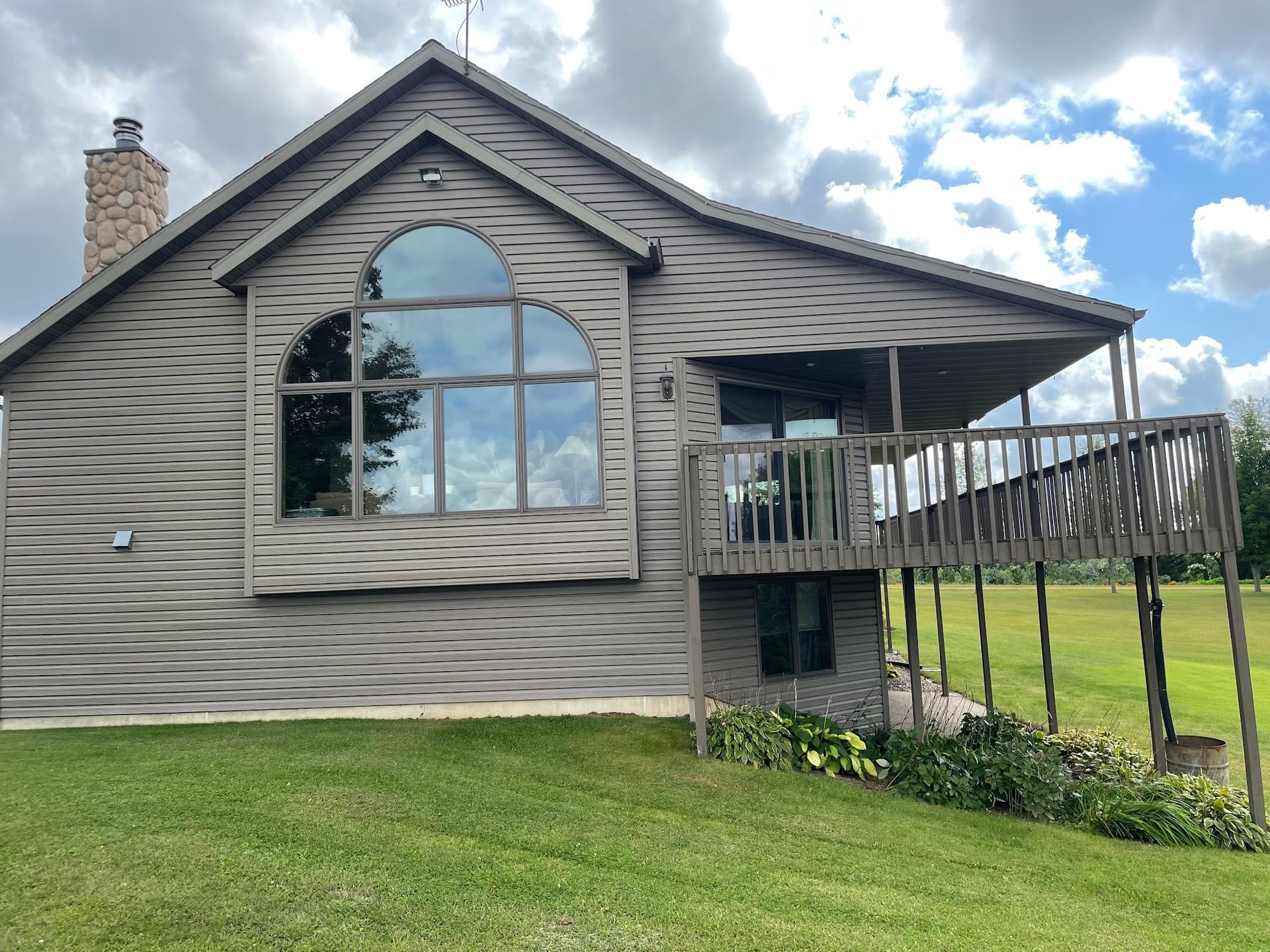 House with brown siding, large arched window, deck, and green lawn under a cloudy sky.
