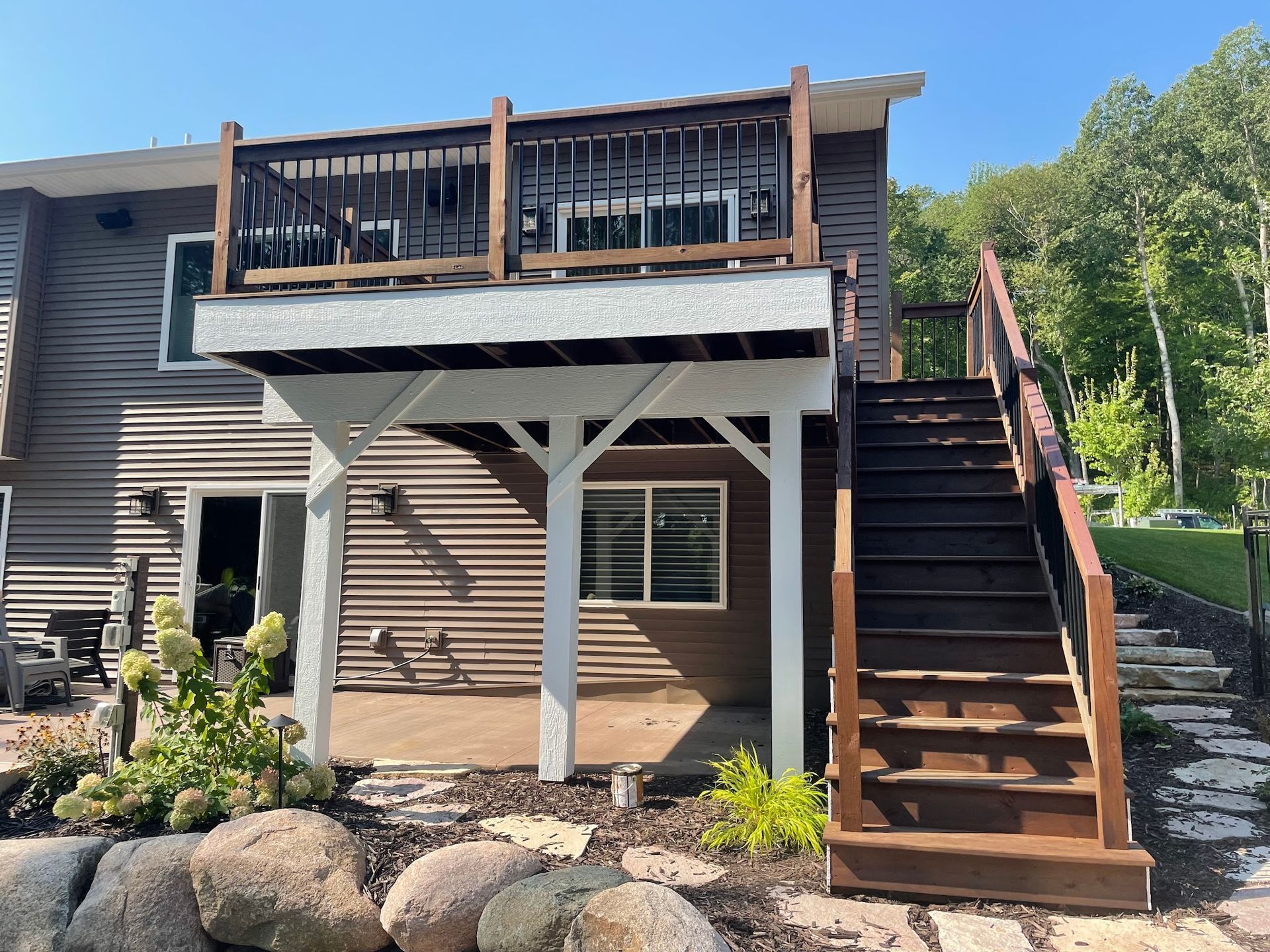 Two-story house with brown siding and deck; stairs lead up to the deck. Rocks and plants in the foreground.