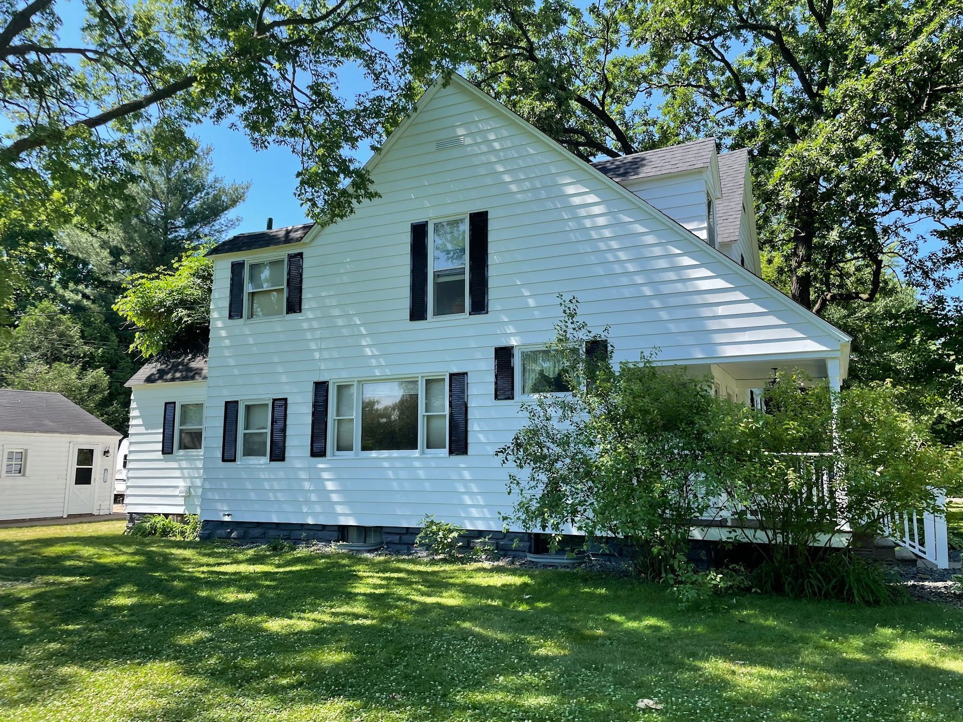 White two-story house with black shutters, surrounded by green grass and trees.