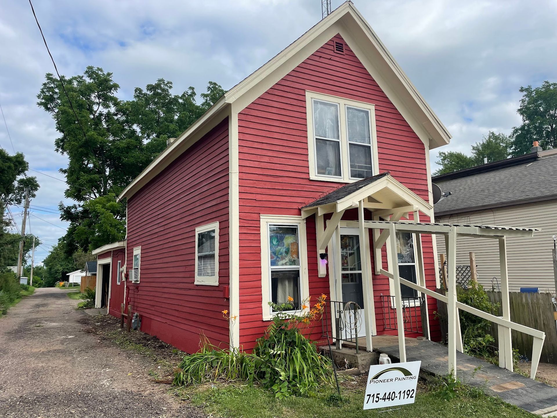 Red two-story house with white trim; overgrown plants and a gravel driveway.