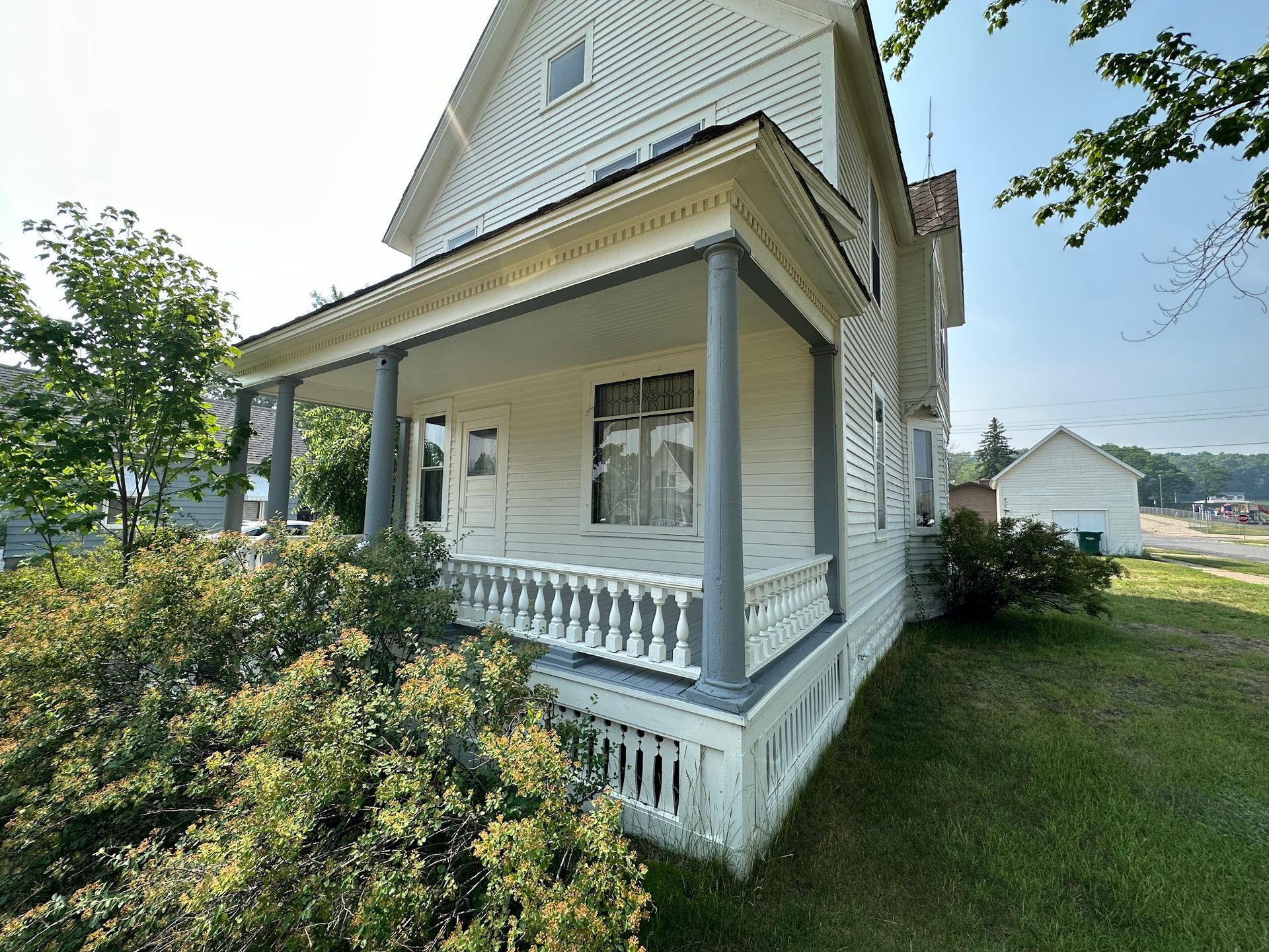White two-story house with porch. Gray columns and trim, bushes in foreground, clear sky.