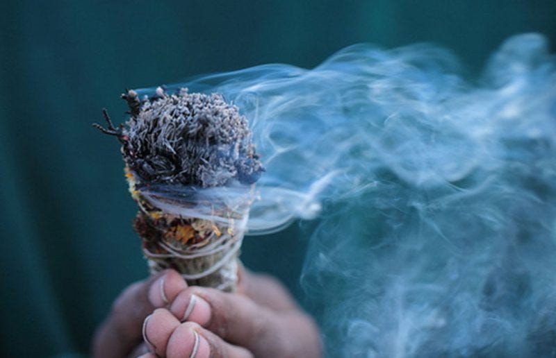 Hand holding a burning sage bundle, emitting white smoke against a teal background.