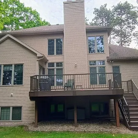 Back of a two-story house with a brown deck, chimney, and stairs. Green grass and trees surround.
