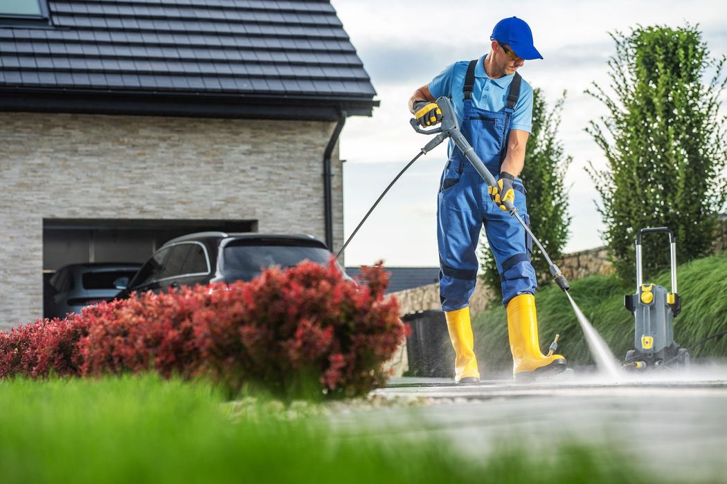 Man power washing a driveway in front of a house; car in garage, red bush and green grass.