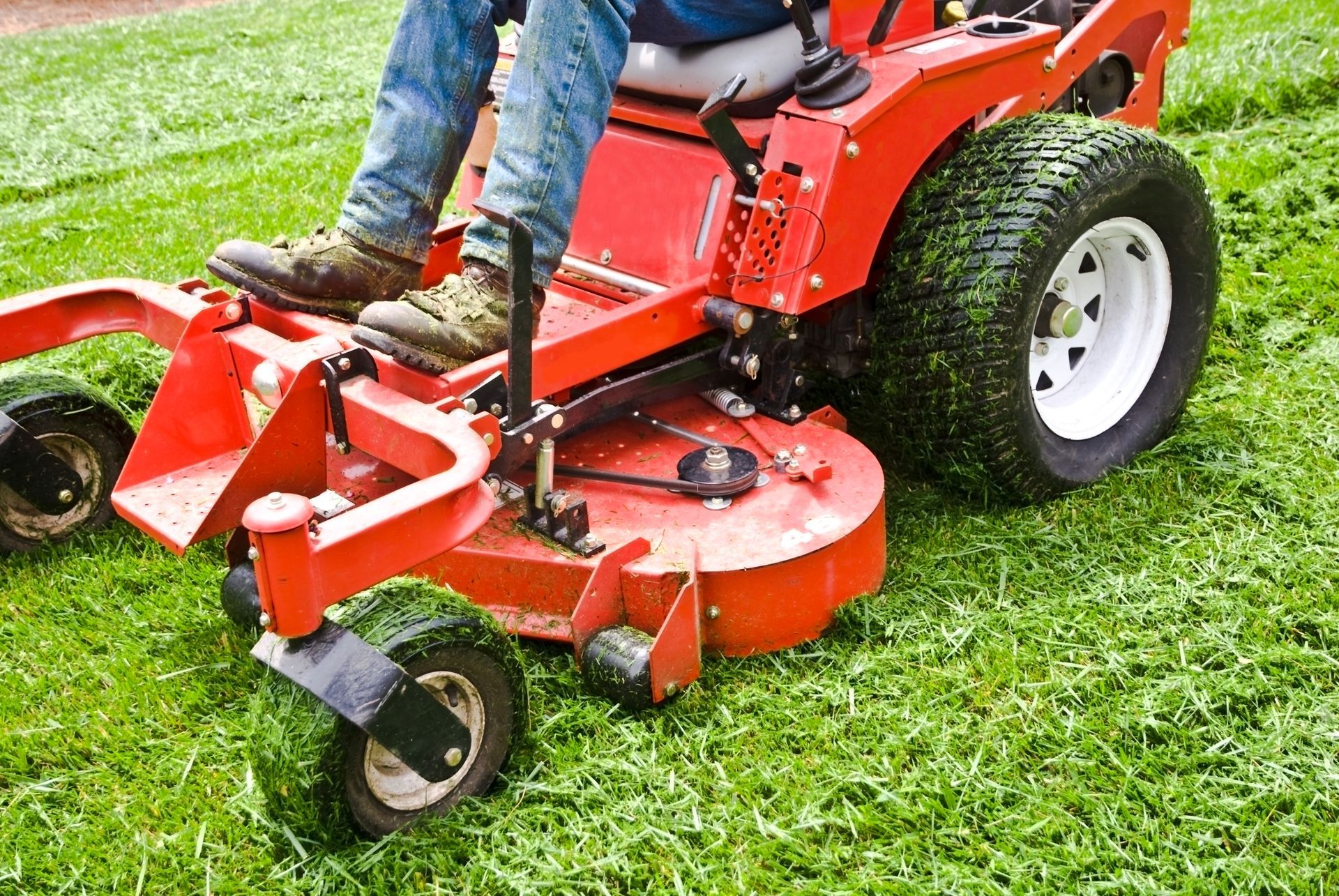 Person operating a red zero-turn lawnmower, mowing a green lawn.