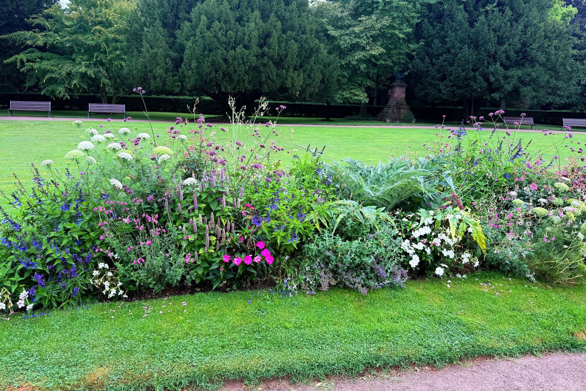 A colorful flower bed borders a green lawn in a park, with trees and benches in the background.