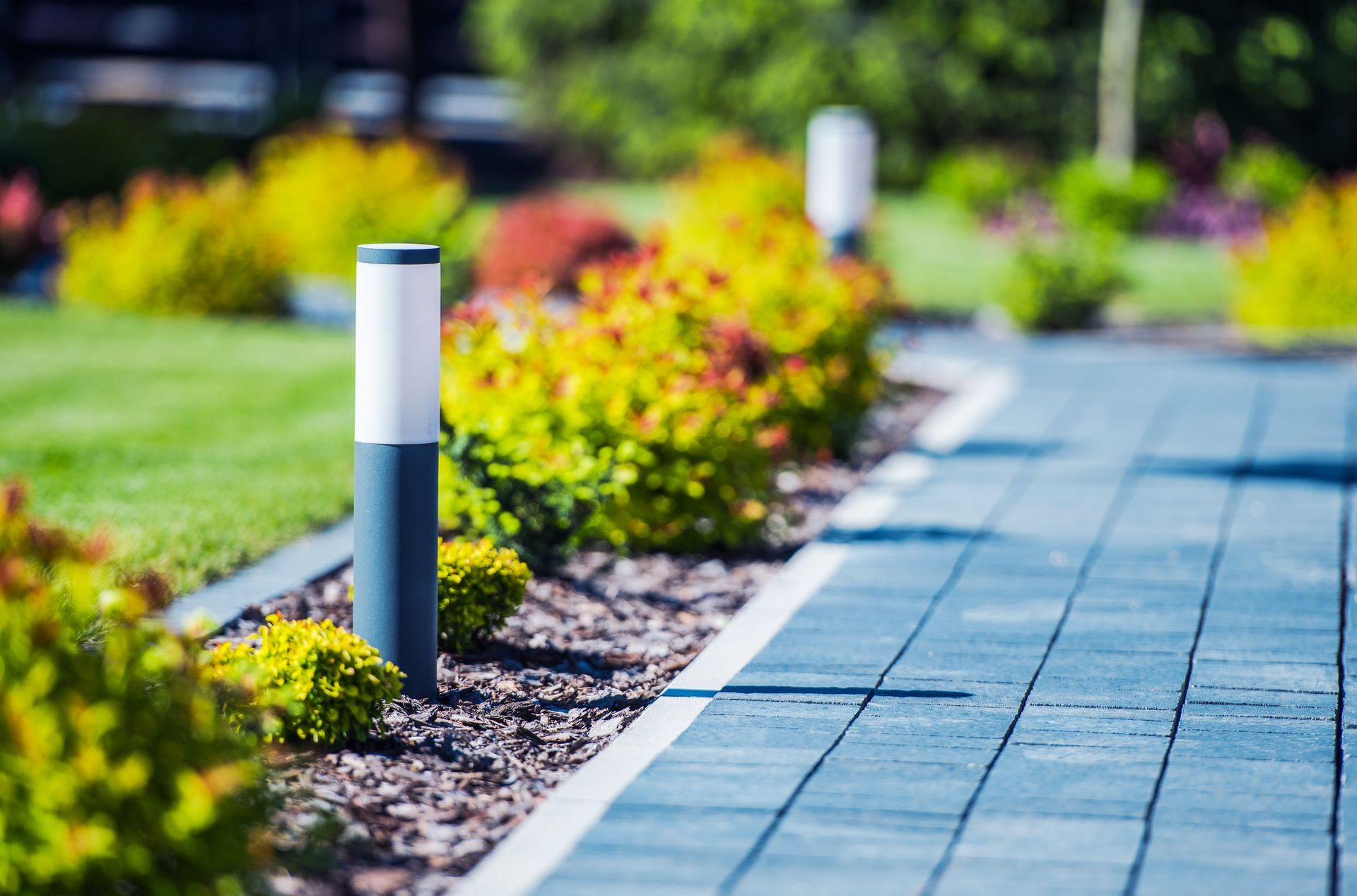 Pathway lined with gray pavers, small green bushes, and modern pillar lights.