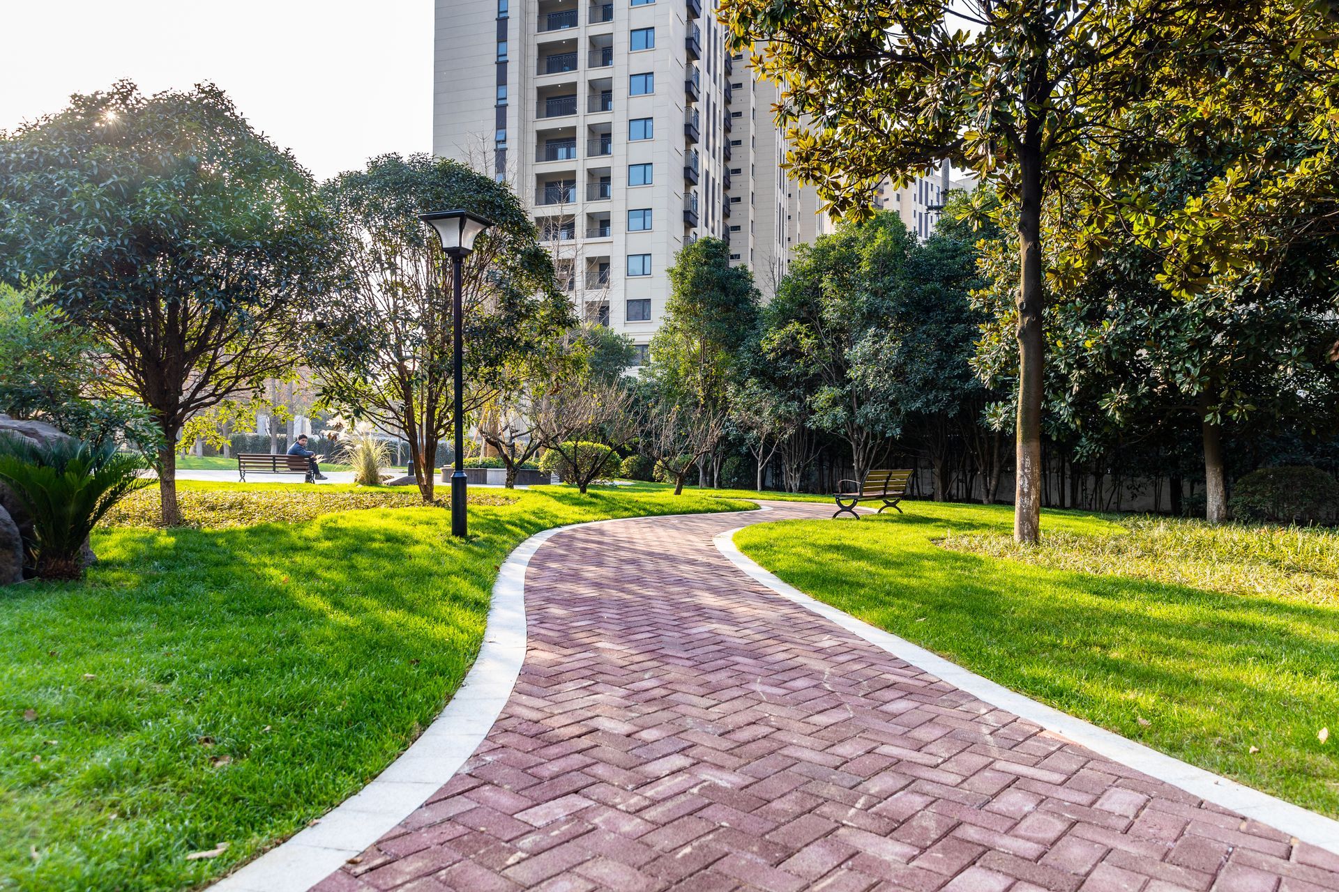 Brick pathway winds through a grassy park, trees line the edges, tall building in the background.