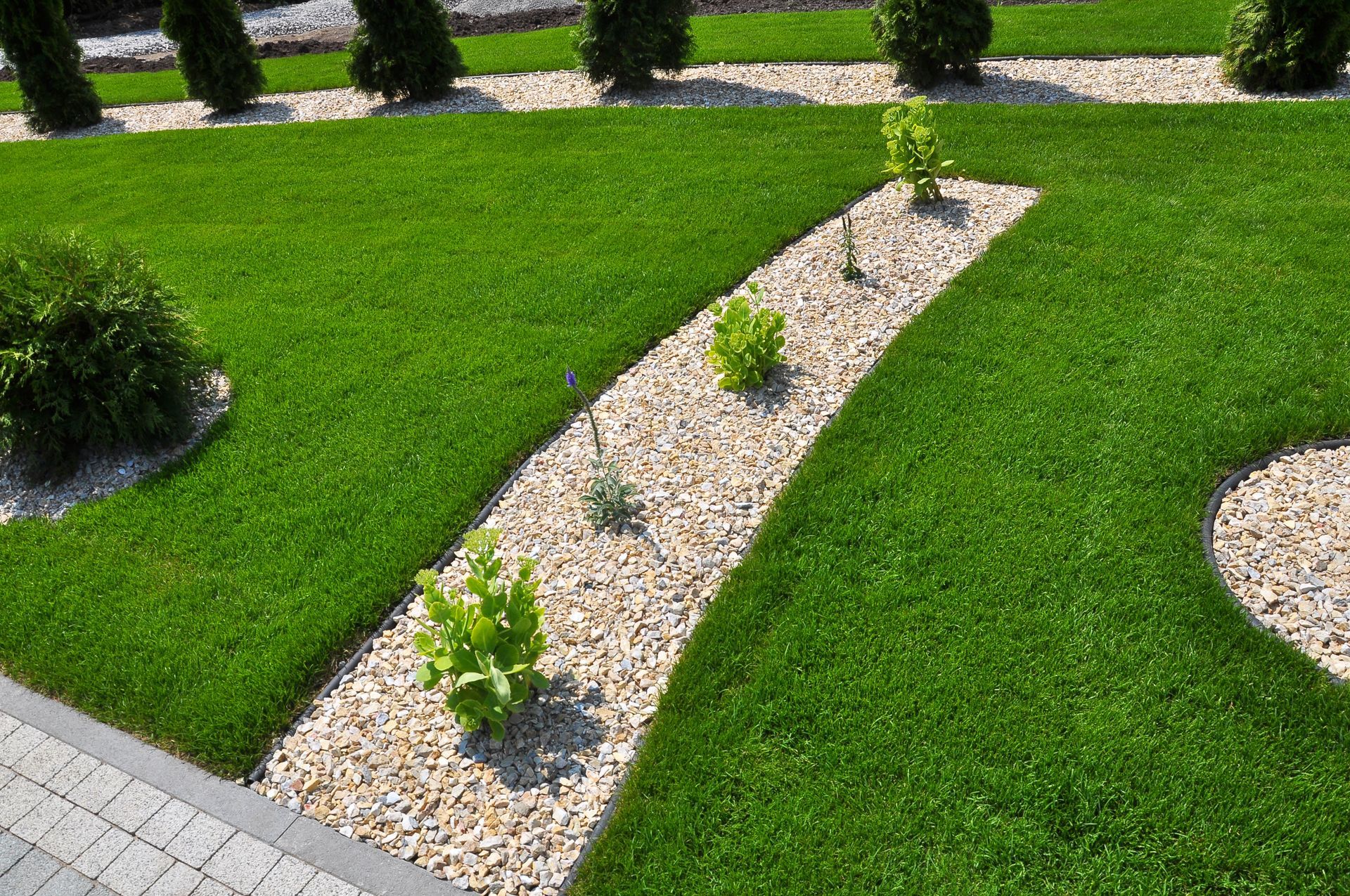 Lush green lawn with a curved gravel bed containing plants. Tall, green trees in the background.