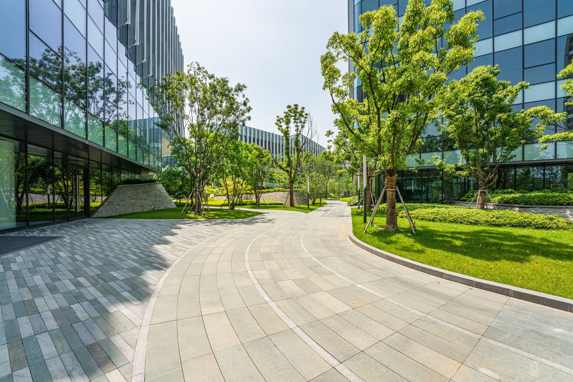 Brick walkway winds through a green, landscaped area between modern glass buildings under a sunny sky.
