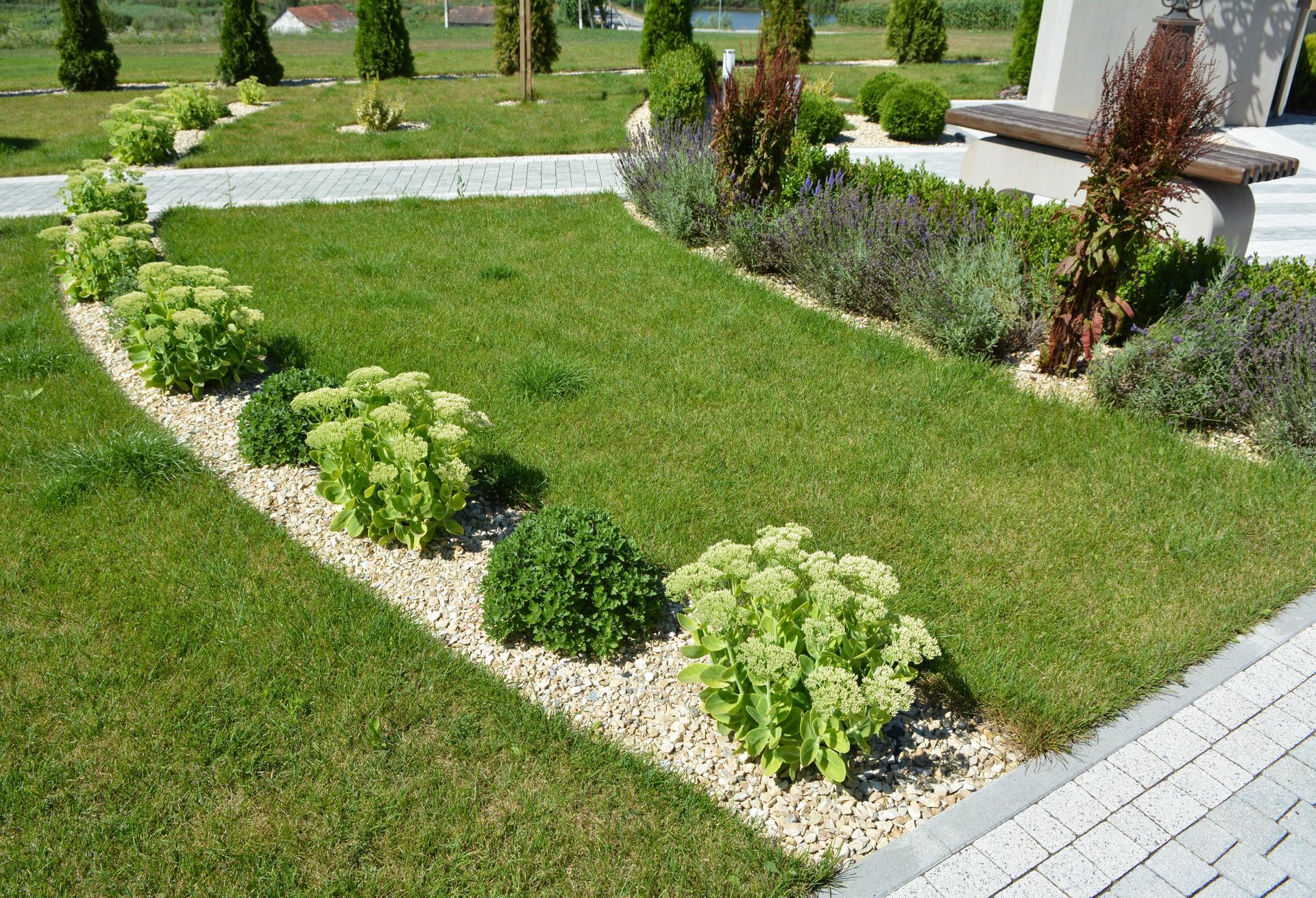 Green lawn bordered by stone and gravel, with a line of green and purple plants.