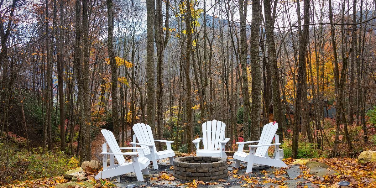 Rustic forest landscape with white Adirondack chairs arranged around a stone fire pit in autumn. Rustic forest landscape with white Adirondack chairs arranged around a stone fire pit in autumn.