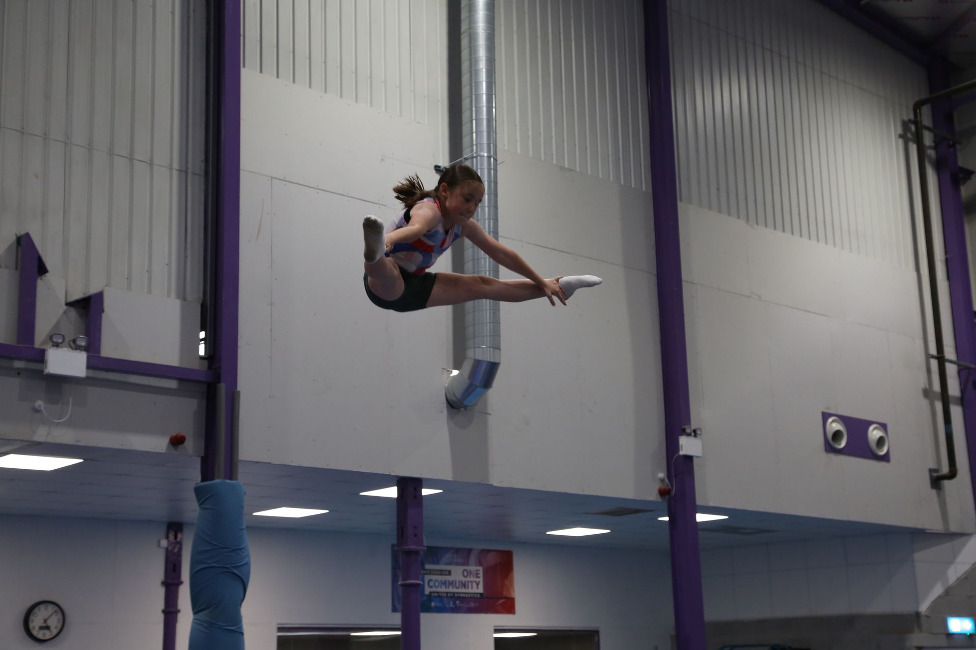 Young athlete performing a split on a trampoline, reaching for a bar in a gym.